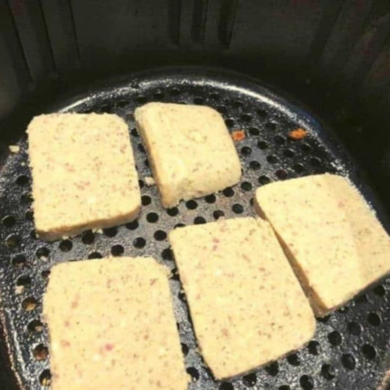 Scrapple slices in an air fryer basket being sprayed with cooking oil