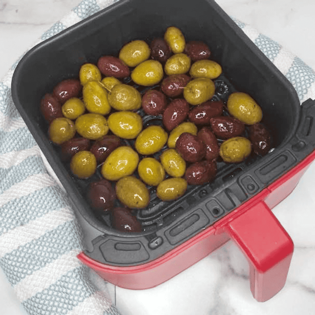 Marinated olives spread evenly in the air fryer basket, ready to cook at 300°F for 5 minutes.