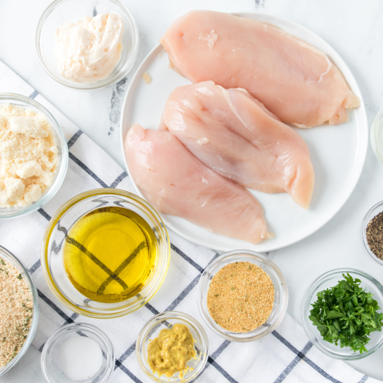Ingredients for chicken costoletta laid out on marble — chicken breasts, panko, lemon, parmesan, eggs, flour, and cream