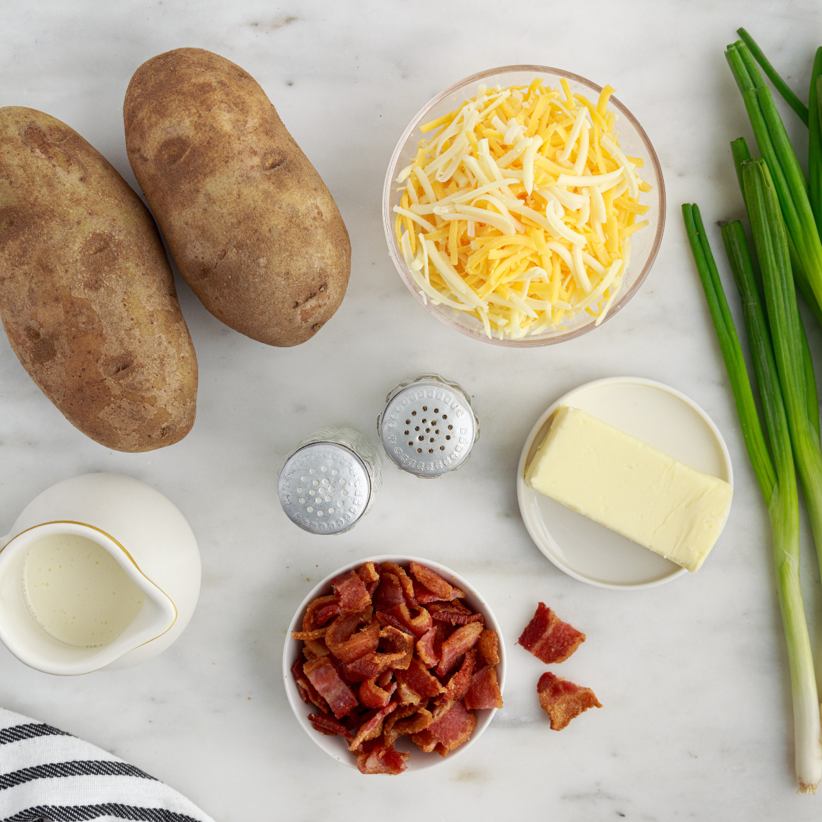 Russet potatoes, butter, sour cream, shredded cheddar, bacon, and chives on a white background