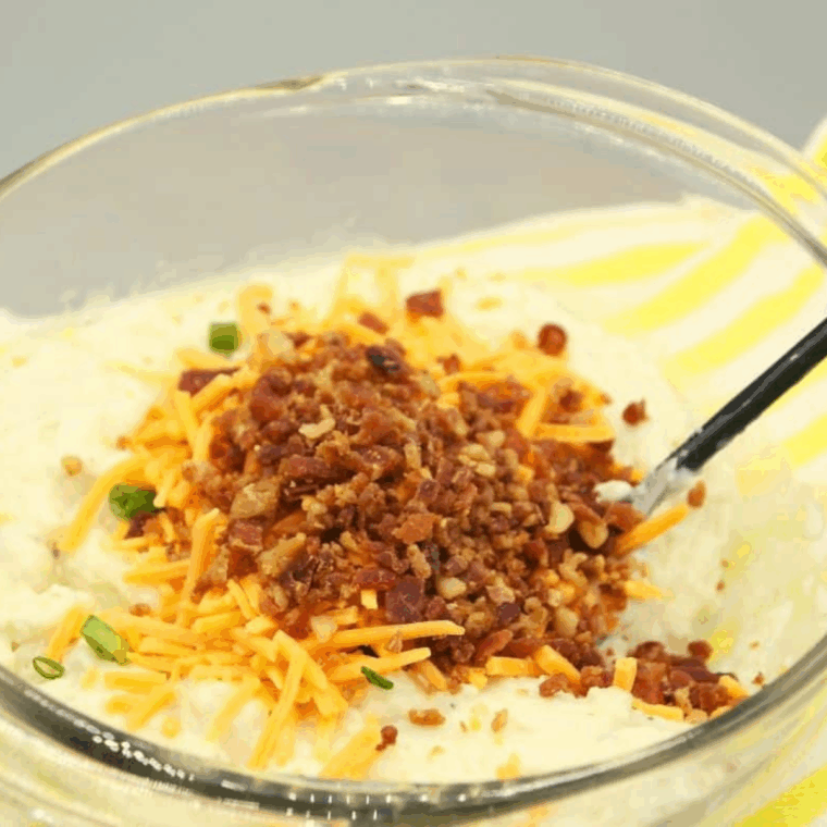 Mashed potatoes, egg, cheddar, bacon, scallion, and flour combined in a glass mixing bowl with a fork.