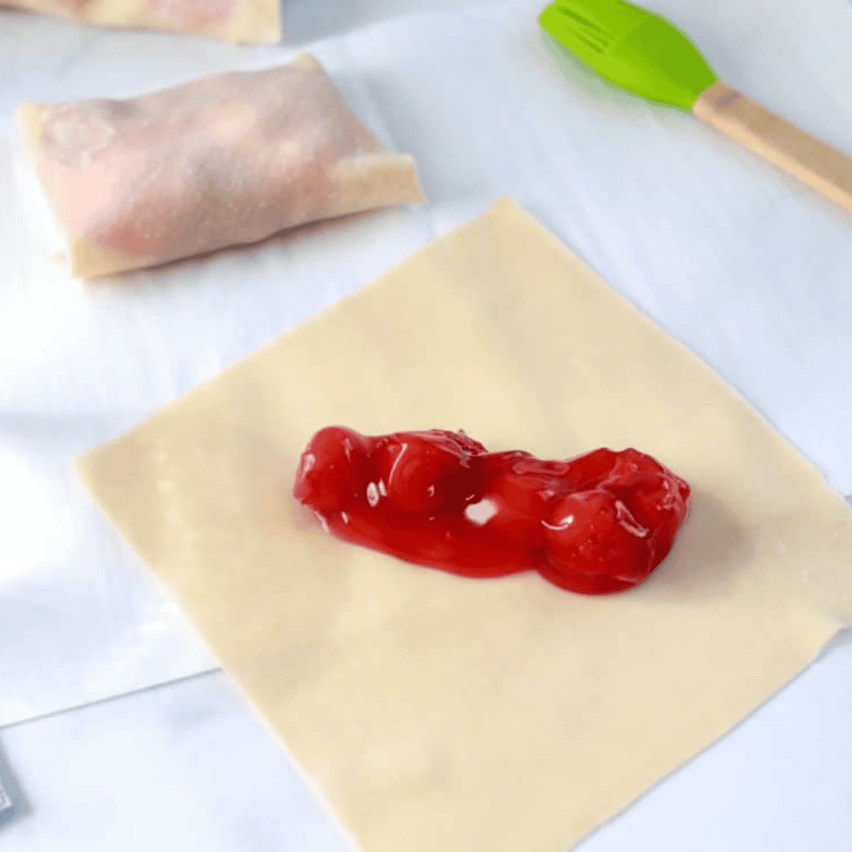 Egg roll wrapper on flat surface with cheesecake mixture and cherry filling placed in the center, ready for rolling.