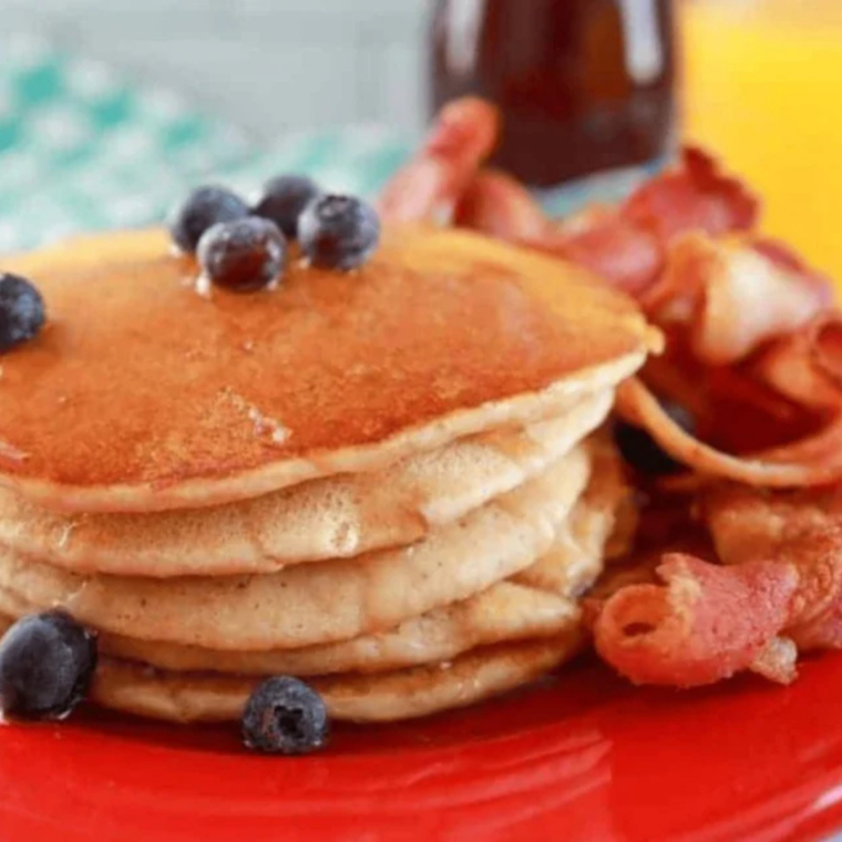 Stack of golden pancakes with butter melting on top, dripping with maple syrup, blueberries scattered around, served on the red plate.