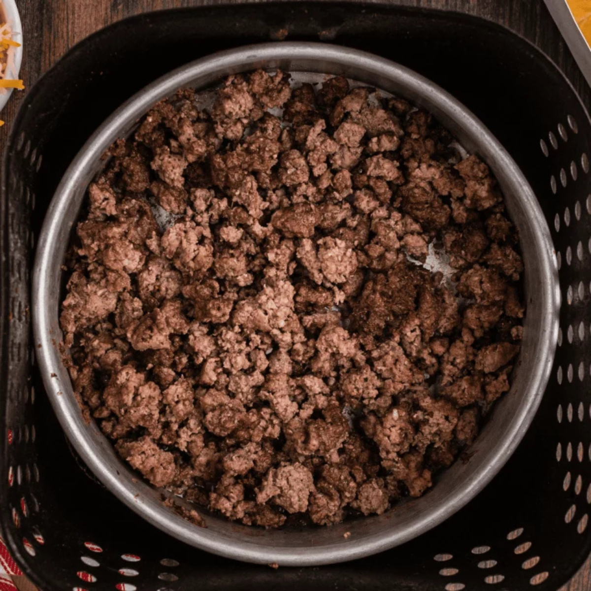 Inserting a meat thermometer into fully browned ground beef in an air fryer basket to ensure it has reached a safe internal temperature of 160°F.