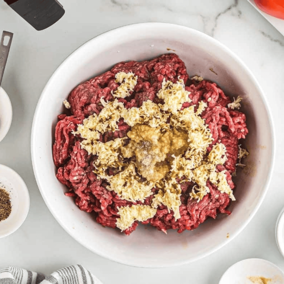 Large white bowl filled with raw beef mince, grated butter, Worcestershire Sauce, garlic, and onion powder.