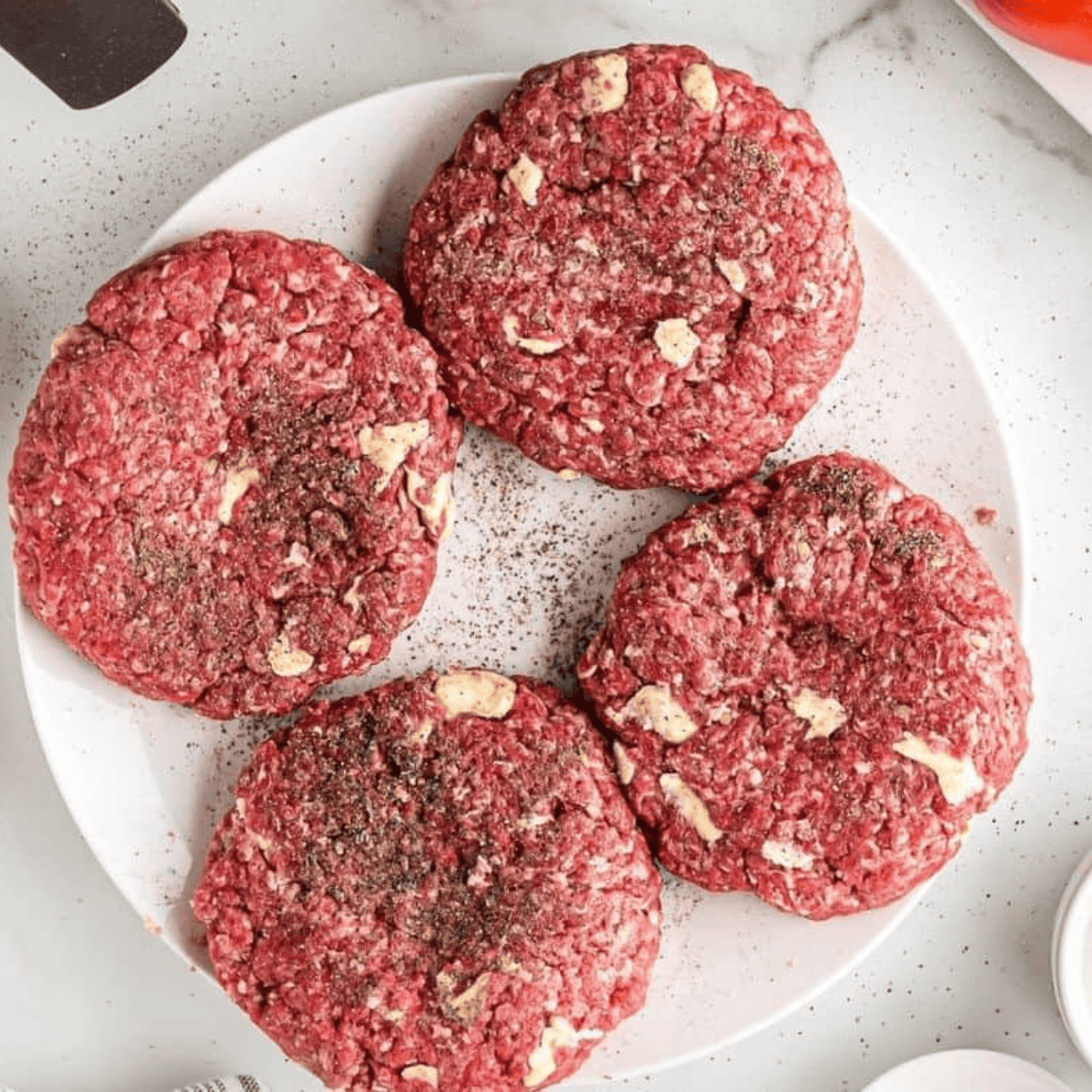 Top view of four raw burger patties on a white plate with salt and pepper sprinkled over them.