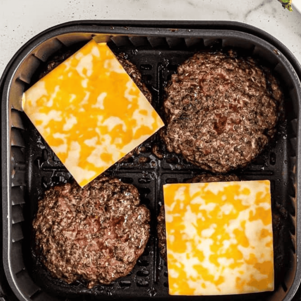 Top view of four burgers in an air fryer, two of them have a square slice of cheese on top.