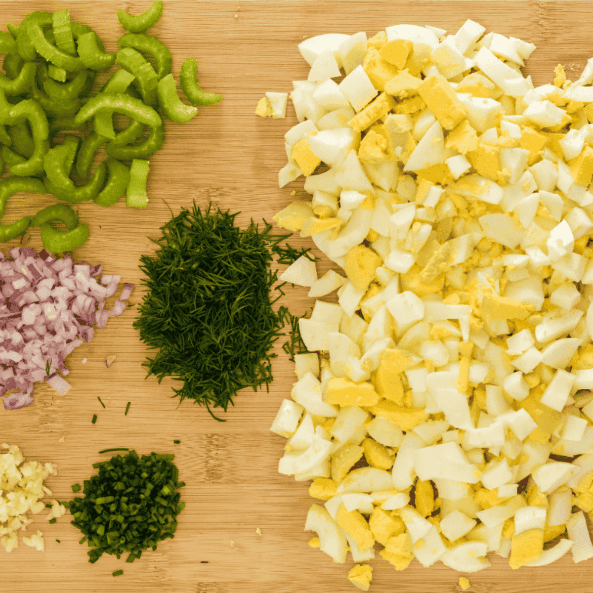 Chopped hard-boiled eggs in a mixing bowl alongside diced dill pickles, celery, and red onions, ready to be combined for egg salad.