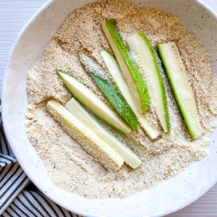 Zucchini stick being dipped in egg wash next to a bowl of almond flour and parmesan coating