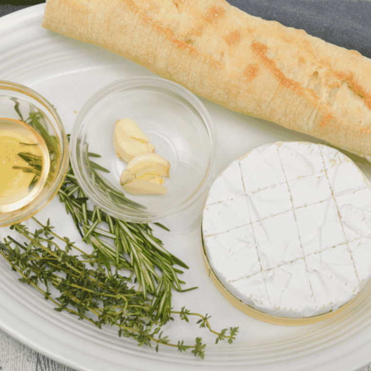 Ingredients for air fryer camembert recipe including cheese wheel, fresh thyme, rosemary, garlic cloves, sea salt, honey, and a baguette
