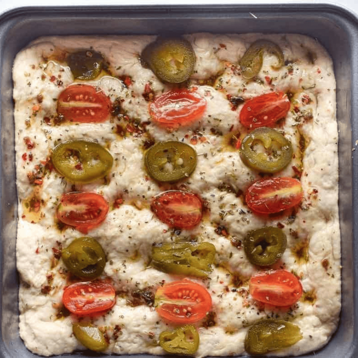 Topping focaccia dough with seasoned olive oil, tomatoes, and jalapenos, then baking until golden brown and cutting into squares.