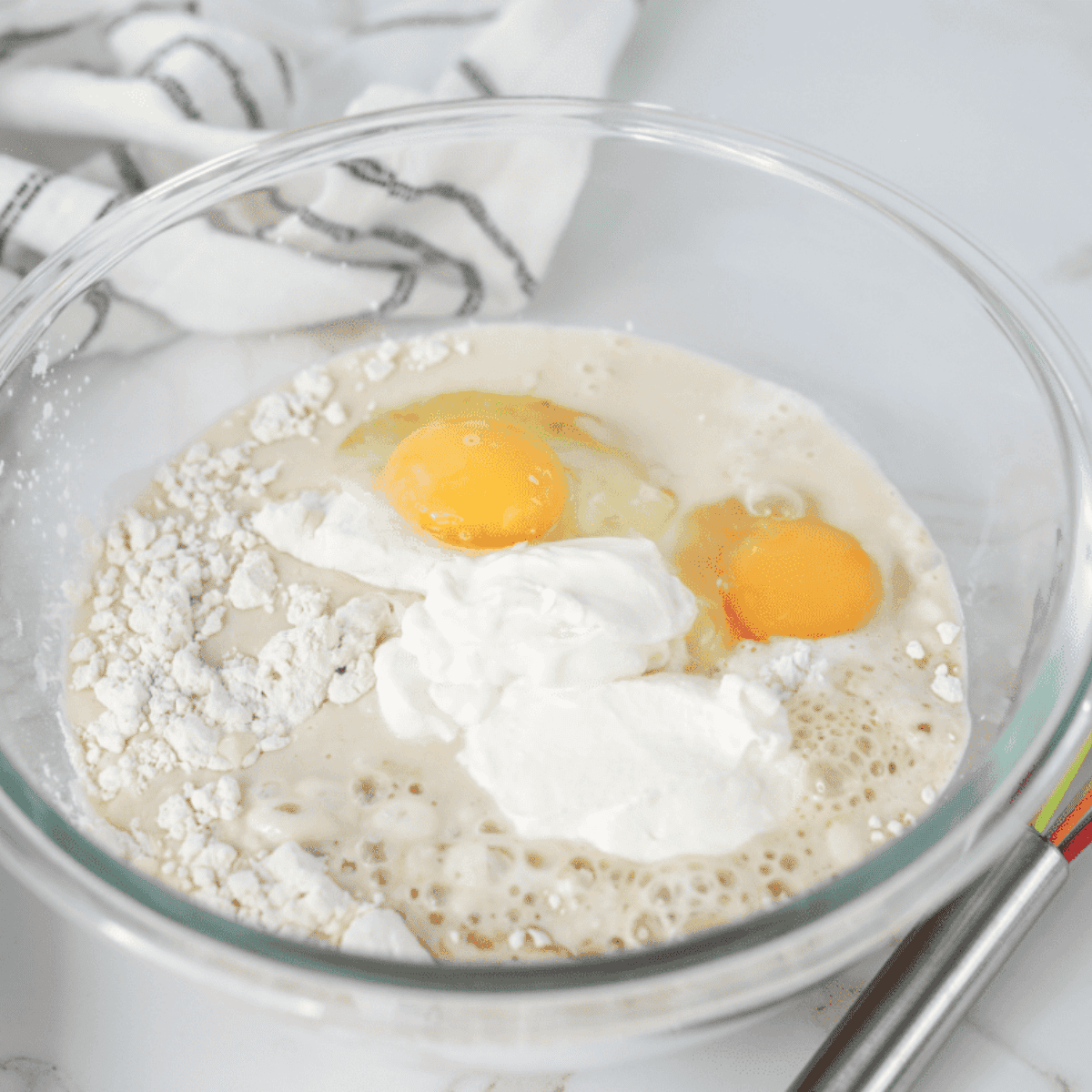Mixing pancake batter ingredients in a large bowl with a whisk, including flour, baking powder, brown sugar, salt, milk, sour cream, eggs, and vanilla extract.