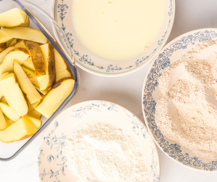 Potato wedges being dredged in seasoned flour with garlic powder and paprika in a shallow bowl