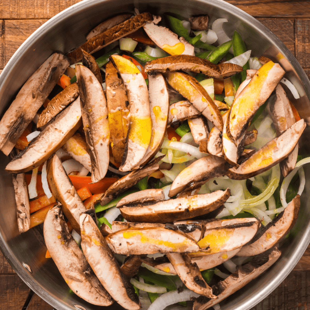 Sliced portobello mushrooms, bell peppers, and onions cooking in a pan with olive oil.