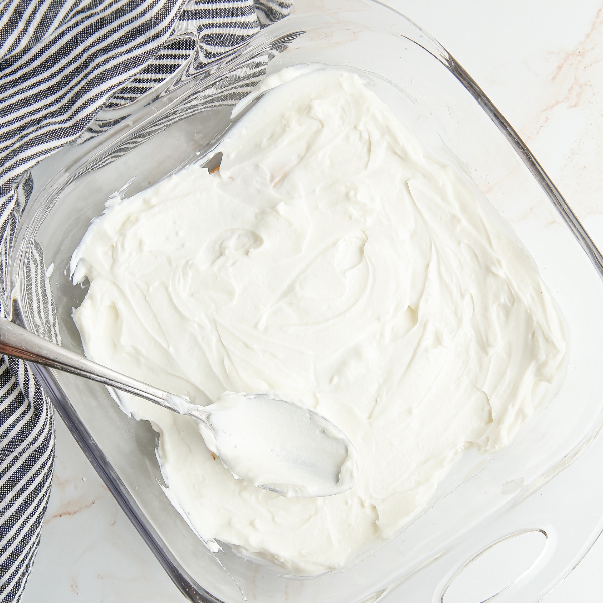Offset spatula spreading mascarpone cream over a layer of espresso-soaked ladyfingers in a square baking dish
