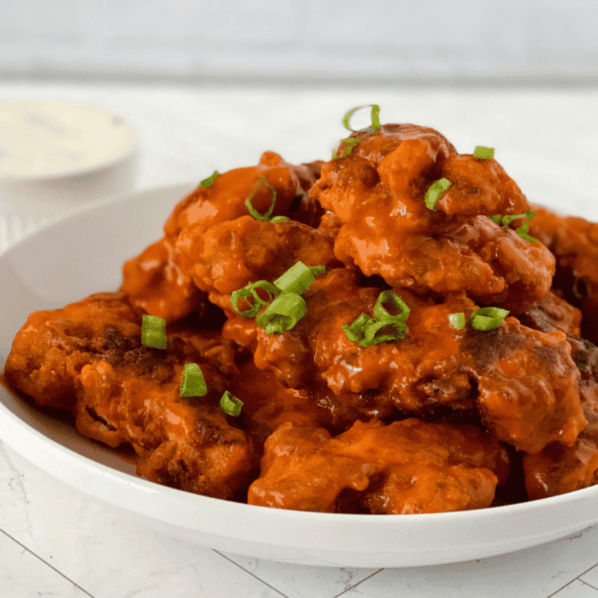 A platter of golden-brown, crispy chicken tenders tossed in a bright orange buffalo sauce, served with a side of celery sticks and a small bowl of creamy blue cheese dressing for dipping.