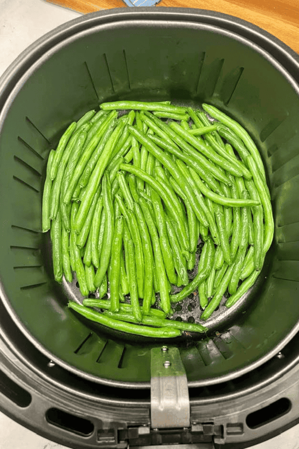 Green beans arranged in a single layer in an air fryer basket, cooking until tender with lightly charred edges.