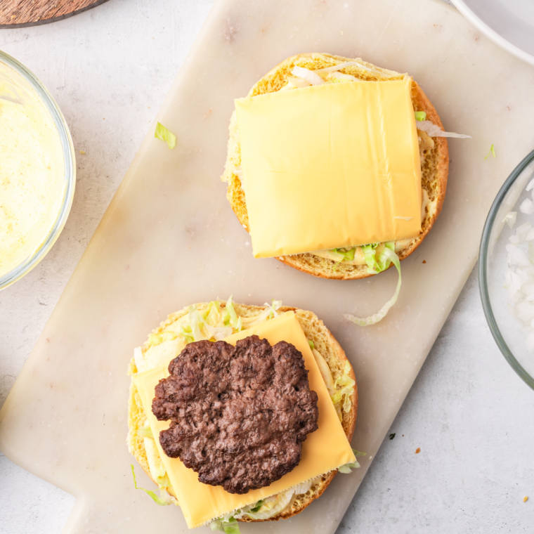 Assembling a homemade Big Mac with layers of special sauce, shredded lettuce, American cheese, beef patties, pickles, and onions between a three-part sesame seed bun.