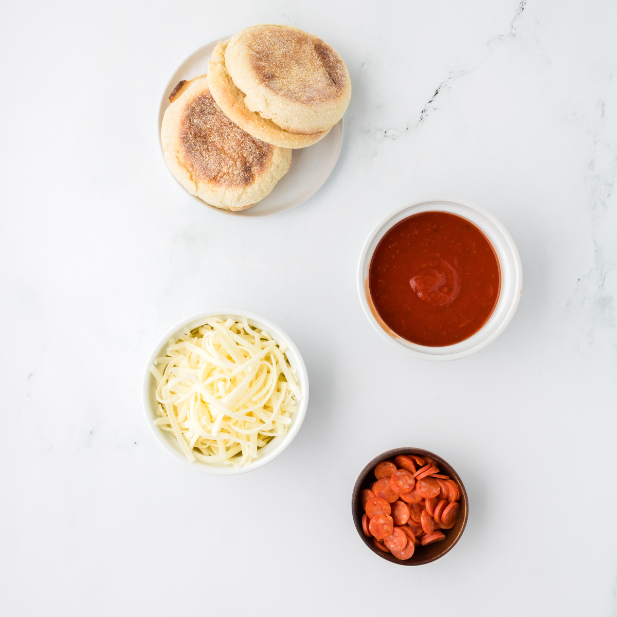 Ingredients laid out for air fryer English muffin pizzas: split English muffins, pizza sauce, shredded mozzarella, mini pepperoni, Italian seasoning, and olive oil
