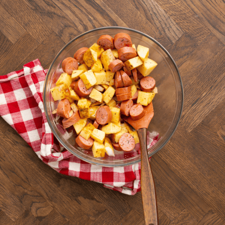Kielbasa and potatoes tossed with olive oil and seasonings in a large bowl.