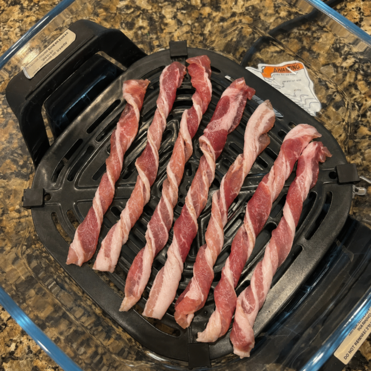 Six raw bacon strips fanned out on parchment beside the Ninja Crispi glass container and crisper plate