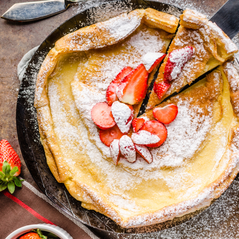 Hero shot of a puffy golden Dutch baby in a small cast iron skillet, dusted with powdered sugar and topped with fresh berries