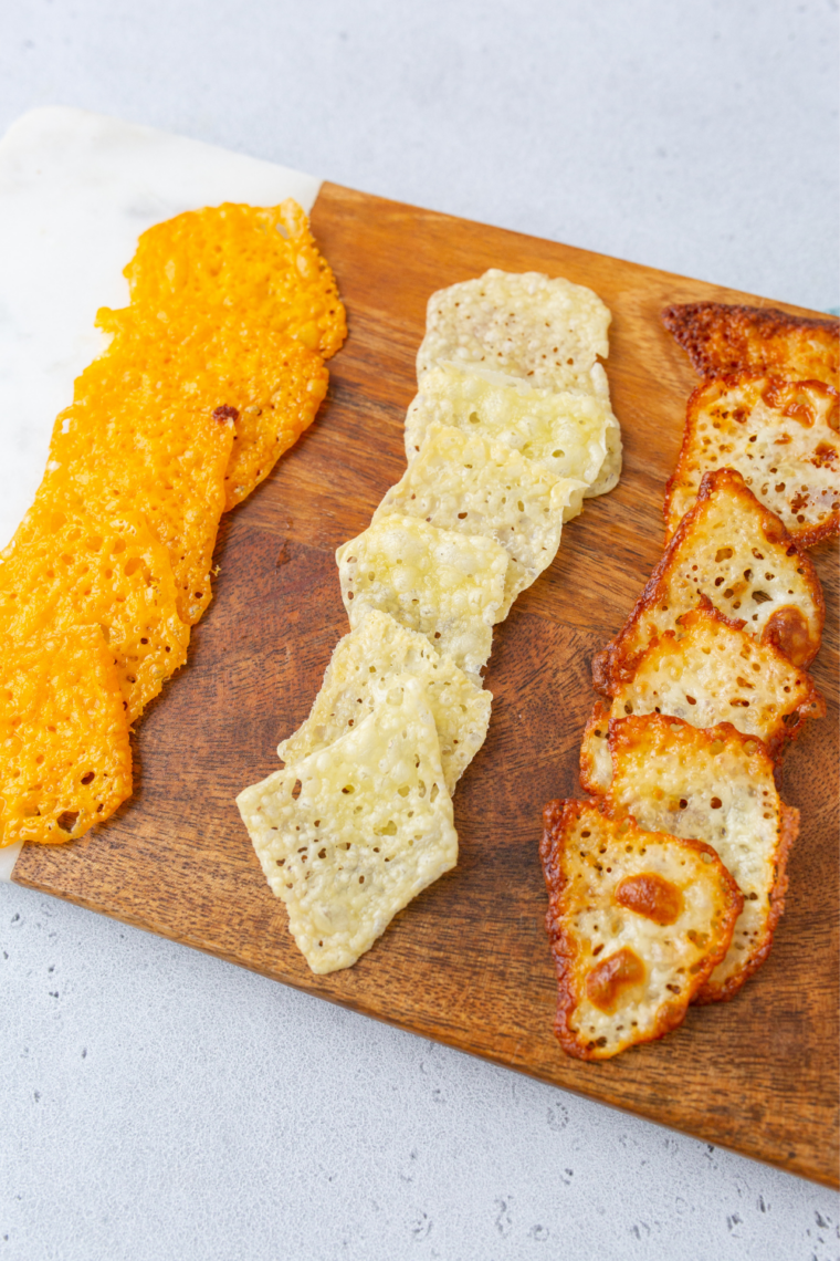Three types of air fryer cheese crisps on a wooden board — cheddar, Parmesan, and pepper jack