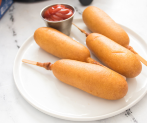 four cooked corn dogs with ketchup on a white plate