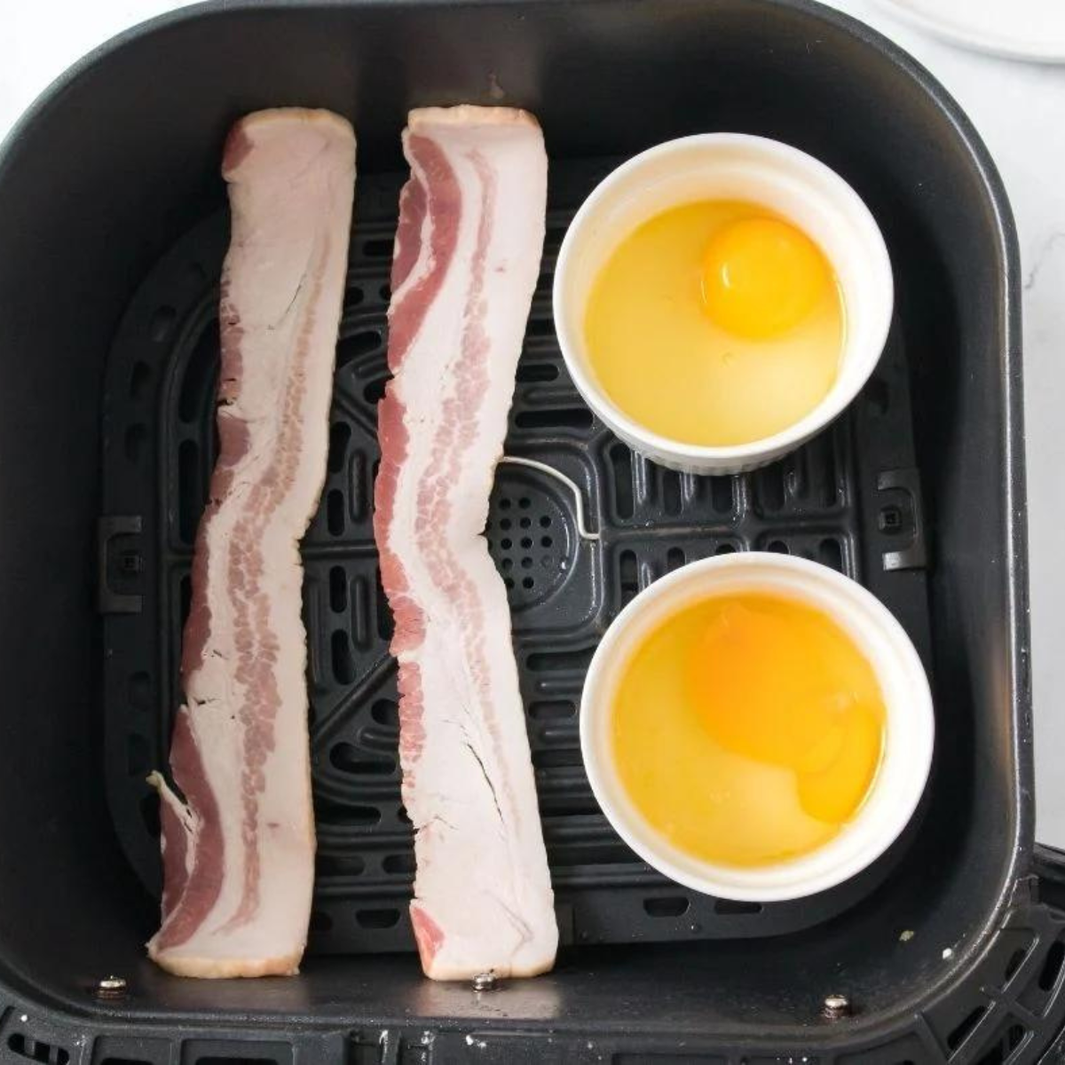 Eggs cooking in a ramekin with sausage and English muffin toasting in the air fryer basket during breakfast sandwich prep.