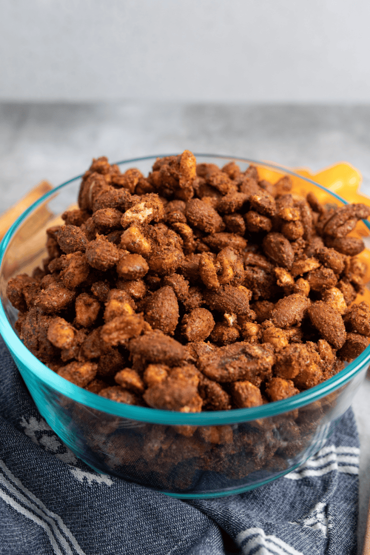 White bowl of herb-roasted mixed nuts on a wooden table beside a scattered handful of almonds and pecans