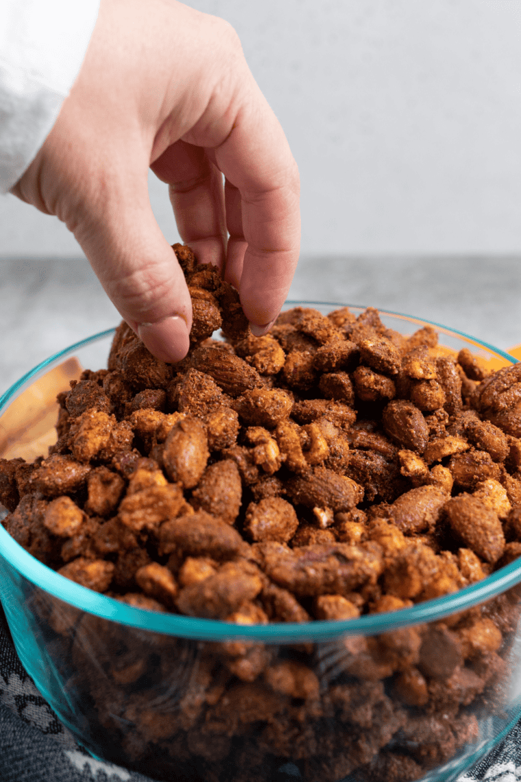 Hand reaching into a white bowl of herb-roasted mixed nuts on a wooden table, almonds and pecans scattered nearby