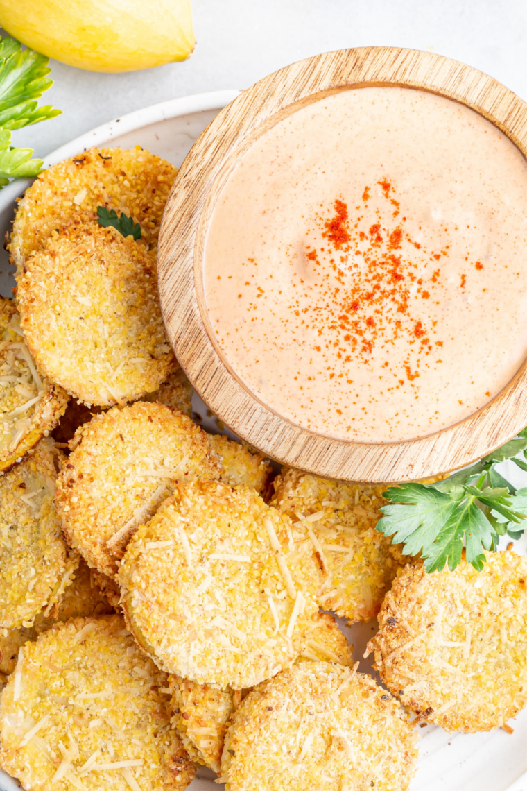 Crispy air fryer summer squash slices on a white plate with a bowl of dipping sauce