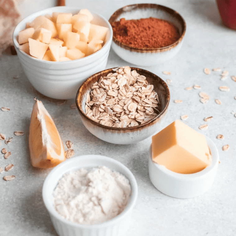 A spread of ingredients for apple crisp, including sliced apples, oats, flour, brown sugar, and cinnamon, arranged on a wooden countertop.