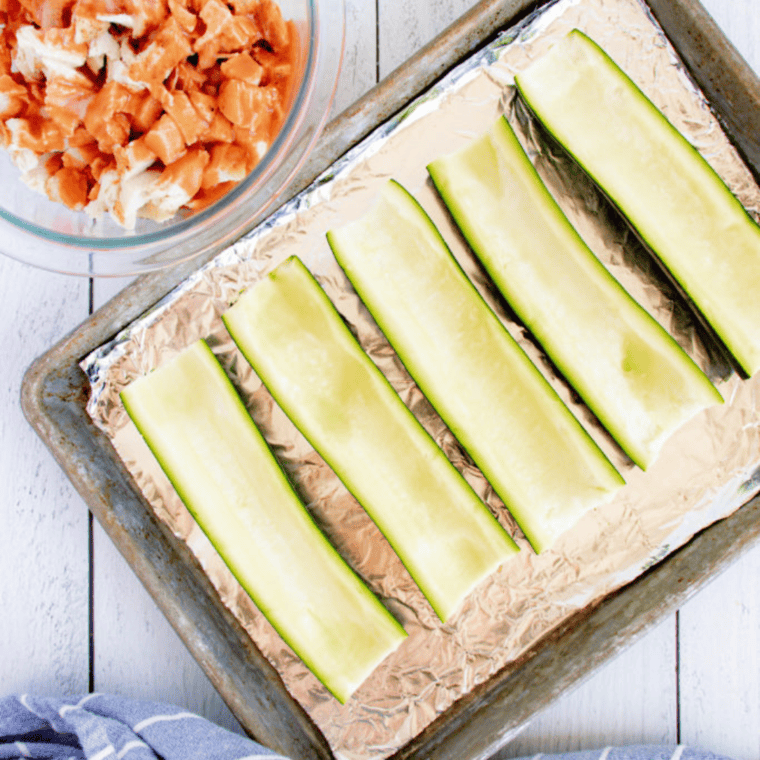 Cutting zucchini in half lengthwise on a cutting board, preparing to scoop out the insides for zucchini boats.