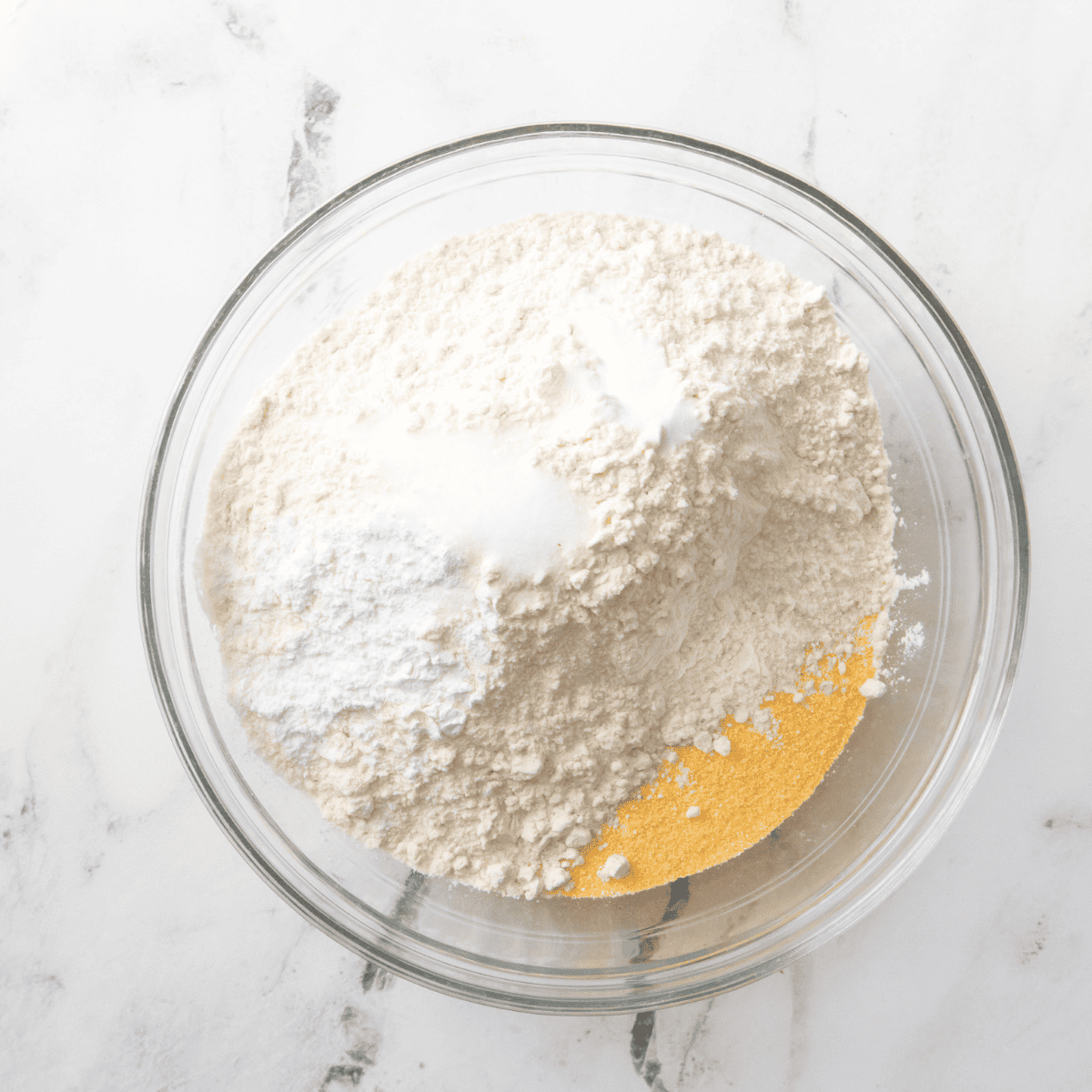 Dry ingredients including cornmeal, flour, sugar, baking powder, and salt in a mixing bowl, with a separate bowl of eggs, milk, and oil beside it.