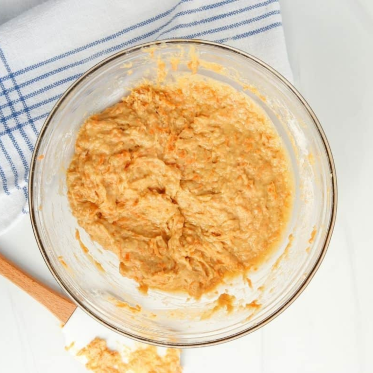 Grated carrots, nuts, raisins, and shredded coconut being folded into carrot cake batter in a mixing bowl, before pouring it evenly into the prepared pan.