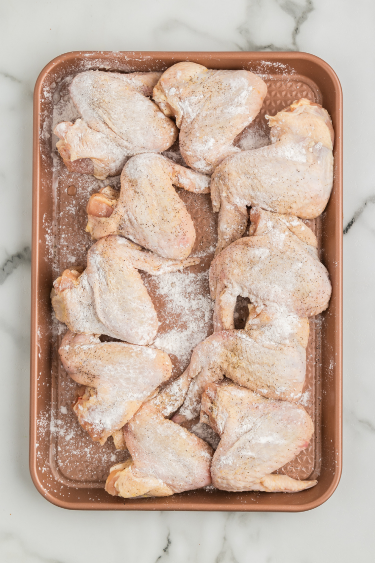 Chicken wings in a mixing bowl being tossed with baking powder, flour, salt, and pepper for a crispy coating.