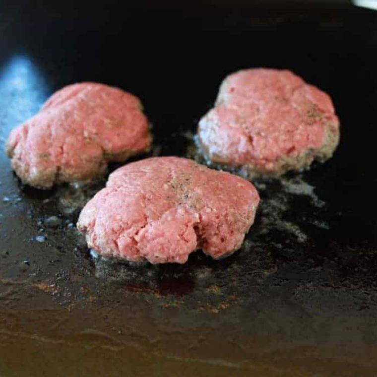 A beef ball being smashed flat on the Blackstone griddle with a burger press through parchment paper.