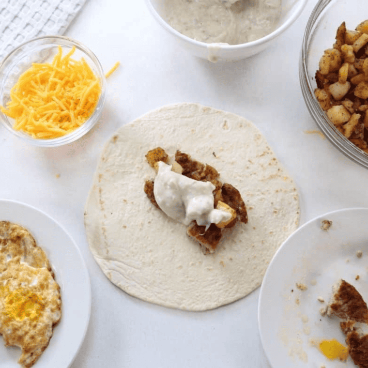 A tortilla topped with diced cooked sausage patties, diced cooked potatoes, and a drizzle of creamy sausage gravy, ready to be wrapped.
