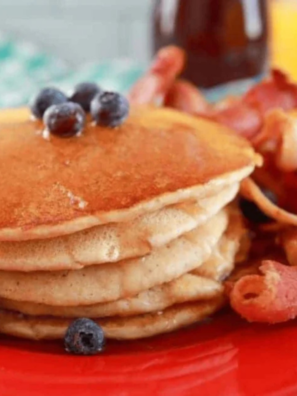 tack of golden pancakes with butter melting on top, dripping with maple syrup, blueberries scattered around, served on the red plate. ]