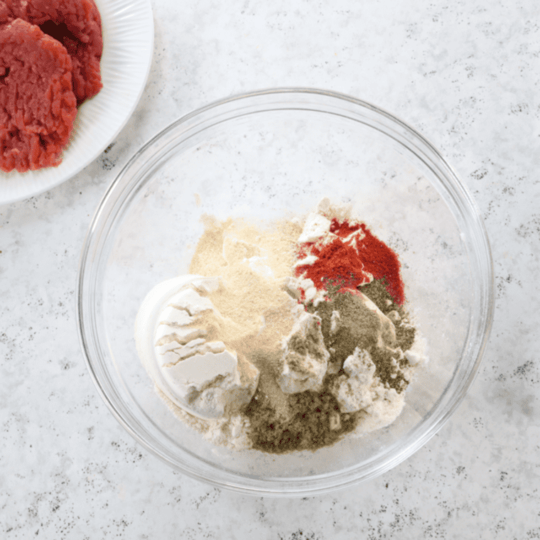 Cube steaks being patted dry with paper towels beside bowls of whisked buttermilk and egg mixture, and a seasoned flour dredge with paprika and spices.