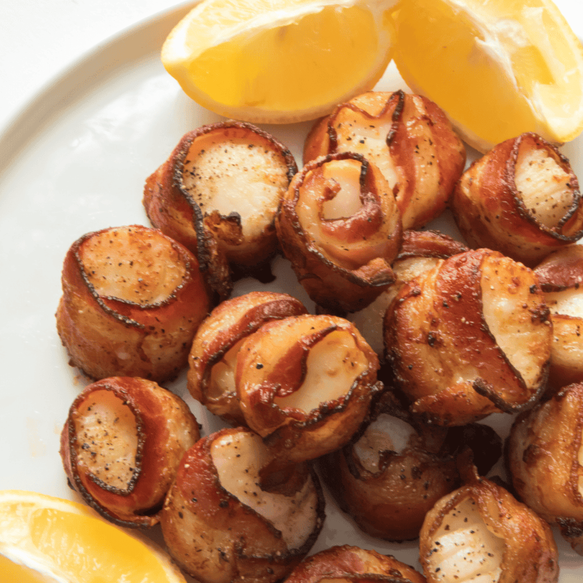 Bacon-wrapped scallops on a white plate with lemon wedges and parsley garnish.