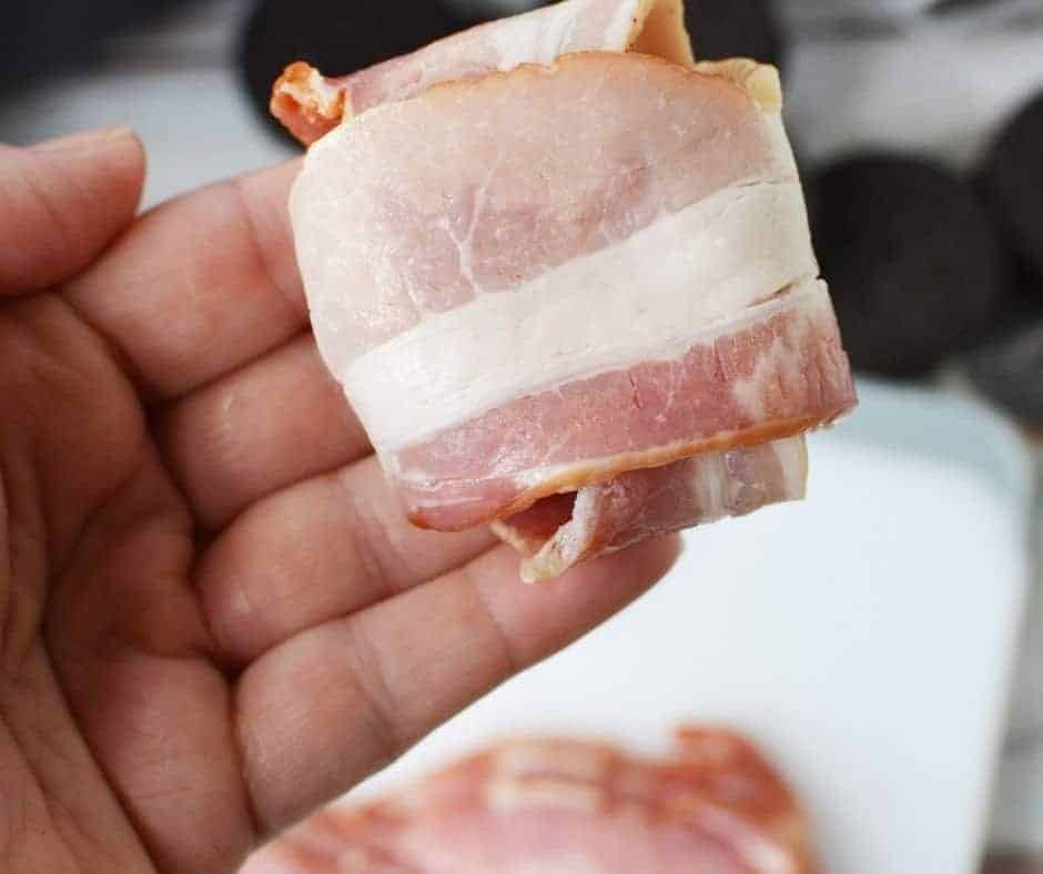 A hand wrapping an Oreo in a slice of bacon, demonstrating the first step of the recipe.