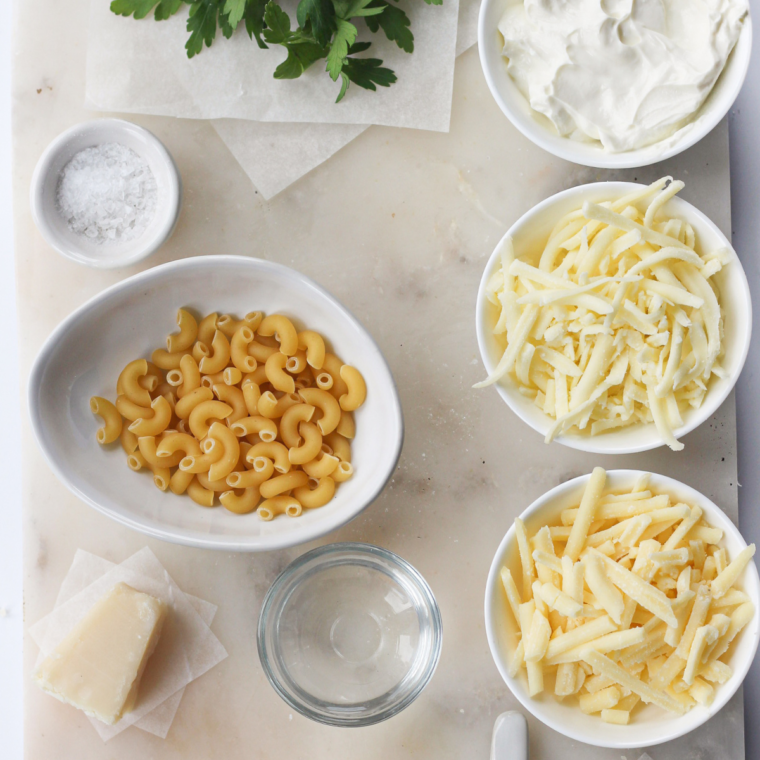 Ingredients for Instant Pot Panera mac and cheese laid out on a wooden countertop, including elbow macaroni, block of sharp white cheddar, American cheese slices, butter, whole milk, Dijon mustard, and hot sauce