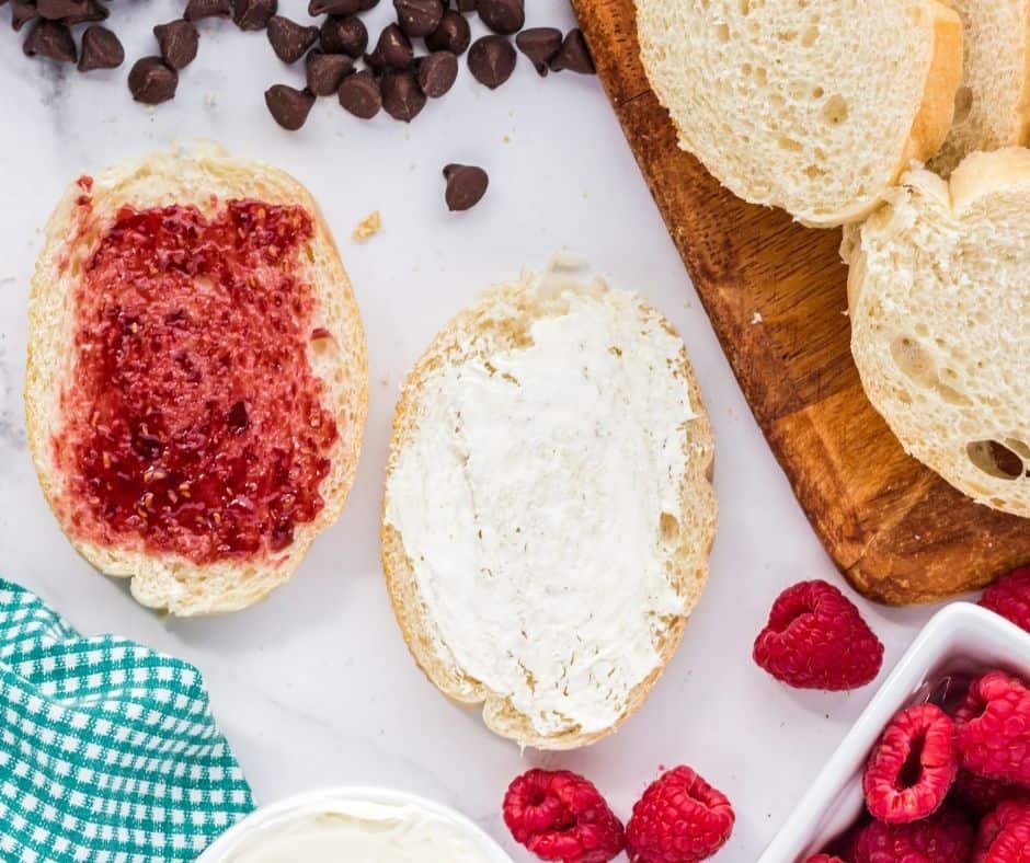 Two slices of French baguette with raspberry preserves and mascarpone cheese spread, ready for sandwich assembly.