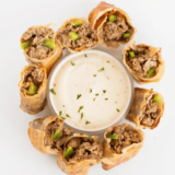 TGI Fridays Philly cheesesteak egg rolls sliced on the diagonal and stacked on a white plate with creamy horseradish dipping sauce