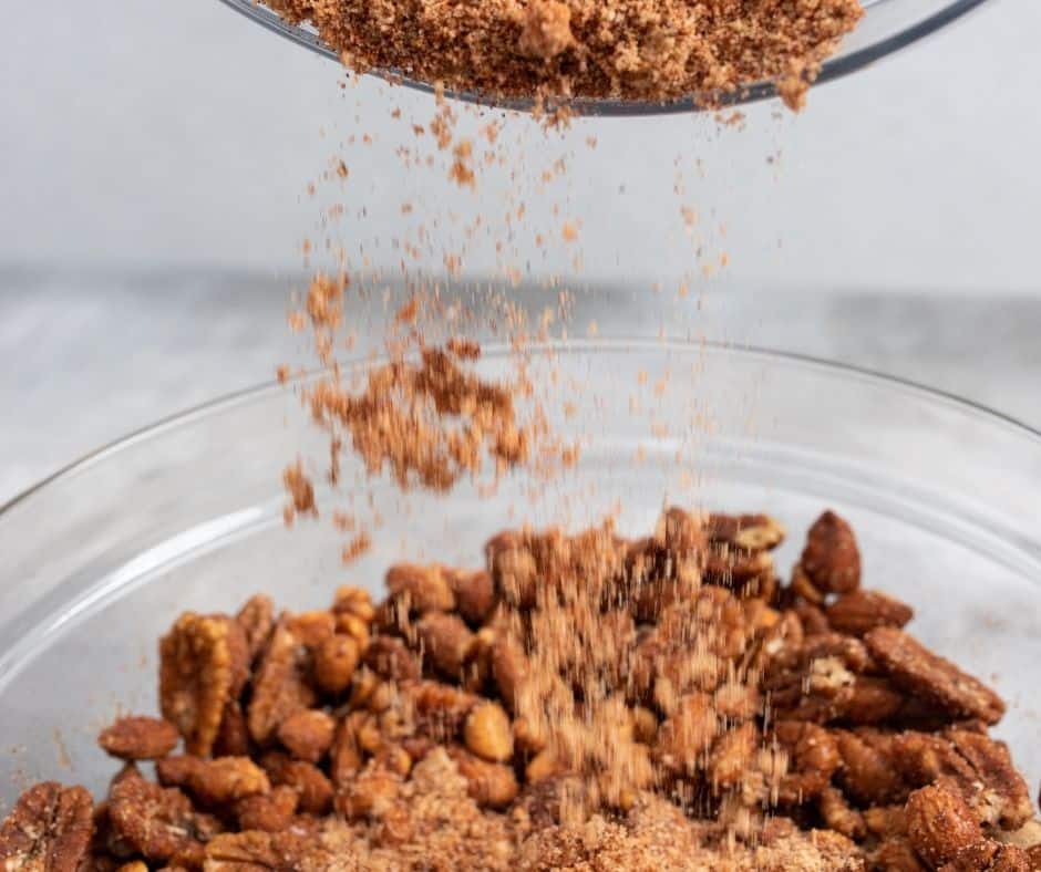 Pecan halves, honey-roasted peanuts, and almonds in a large bowl, coated with frothy egg white and topped with spice mixture, ready to be mixed.