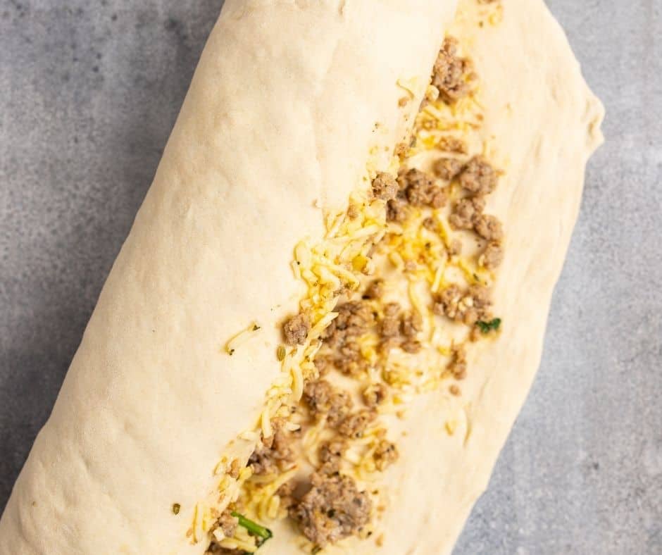 Roll out dough on a floured surface into a rectangle shape, then add sausage filling in the center, leaving a border, and roll it up.