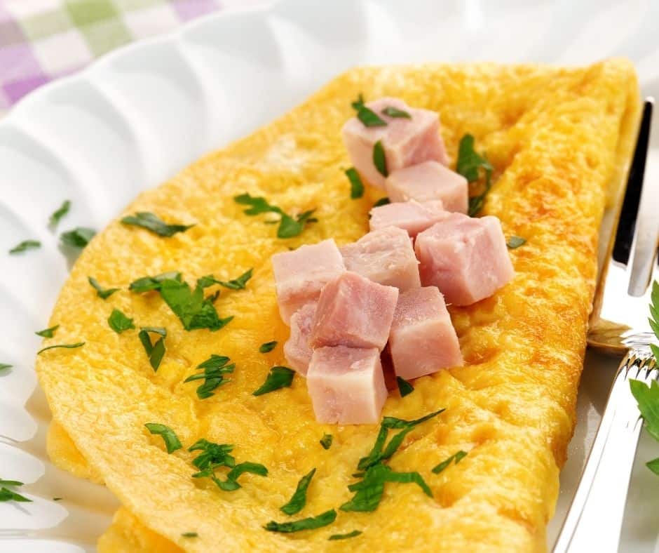 Finished air fryer ham and cheese omelette folded in half on a white plate, topped with fresh chopped chives and a crack of black pepper.