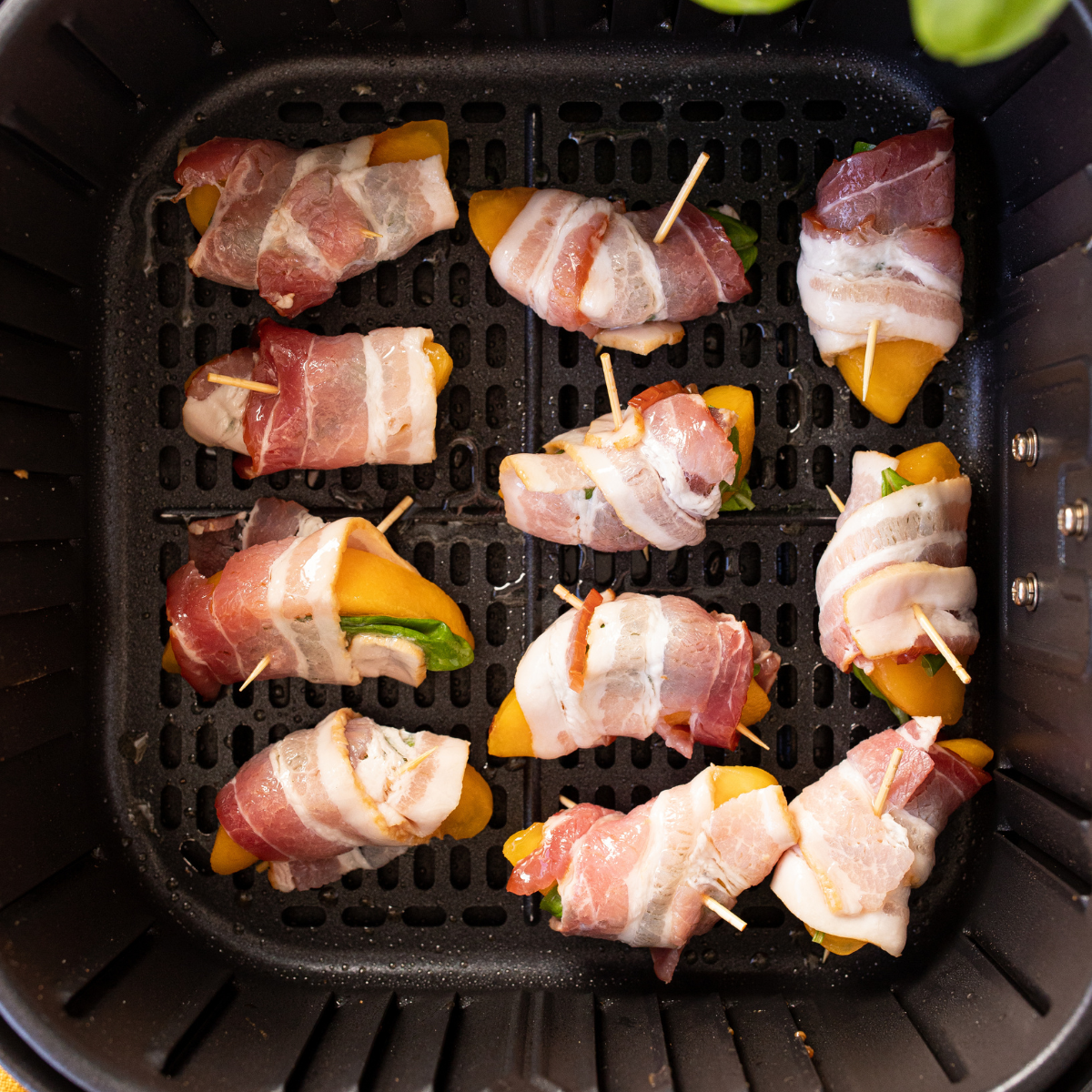 Bacon-wrapped peaches placed in an air fryer basket, brushed with olive oil, ready to cook at 370Β°F for 10-12 minutes.
